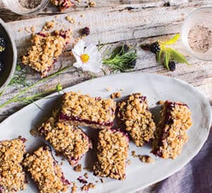 Blackberry oat crumble bars on a white oval plate laid on a butcher block with bowl of fresh blackberries and blackberry flowers