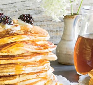 9 layer stack of pancakes topped with butter, syrup and blackberries and set on a table with pitcher of syrup and vase of flowers