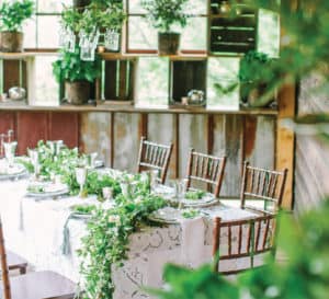 Tablescape of white table cloths, bamboo chairs, and large green swag cascading off the table