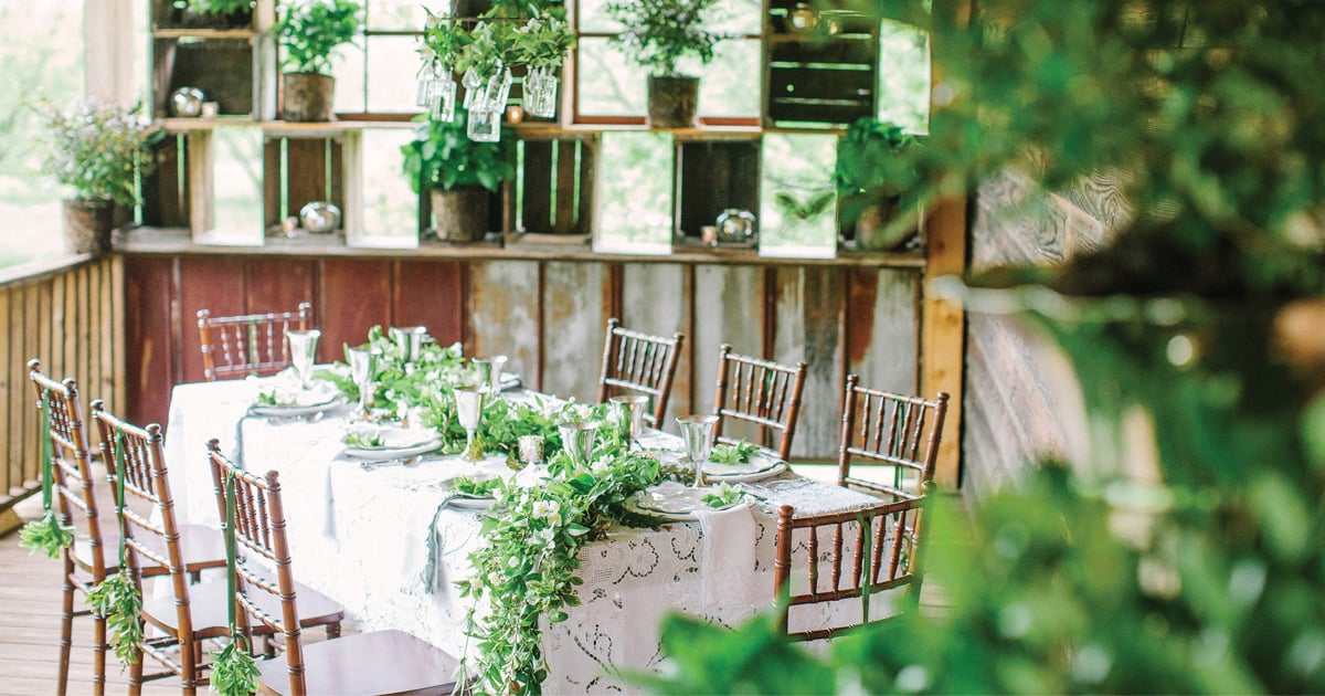 Tablescape of white table cloths, bamboo chairs, and large green swag cascading off the table