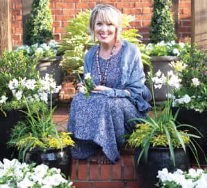 Linda Vater in a blue print dress sits on a brick pathway among Southern Living plants