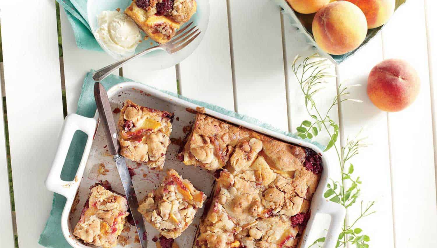 On a white picnic table sits peaches alongside a baking dish of peach cobbler